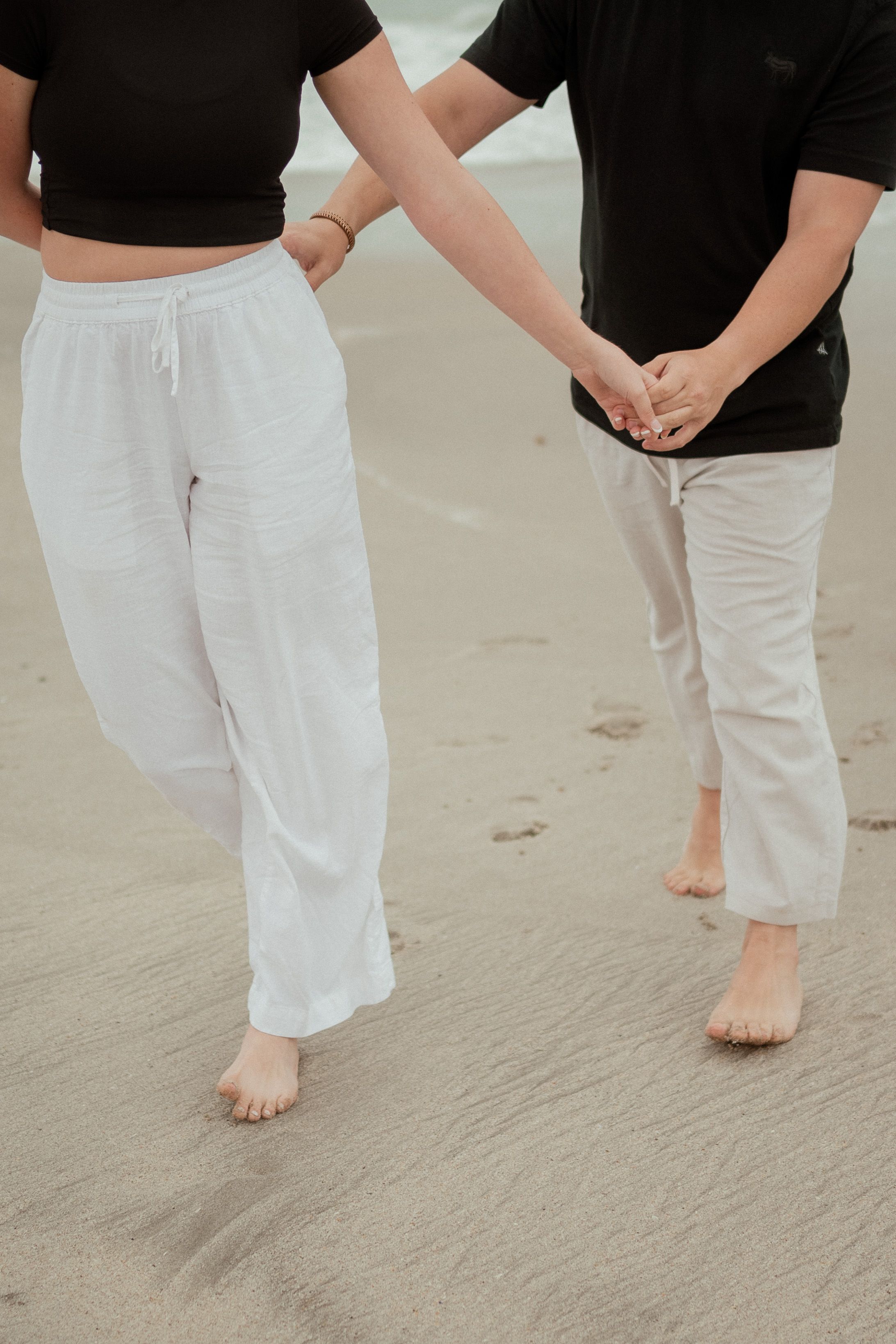 Two people holding hands while walking barefoot on sandy beach wearing casual clothes.