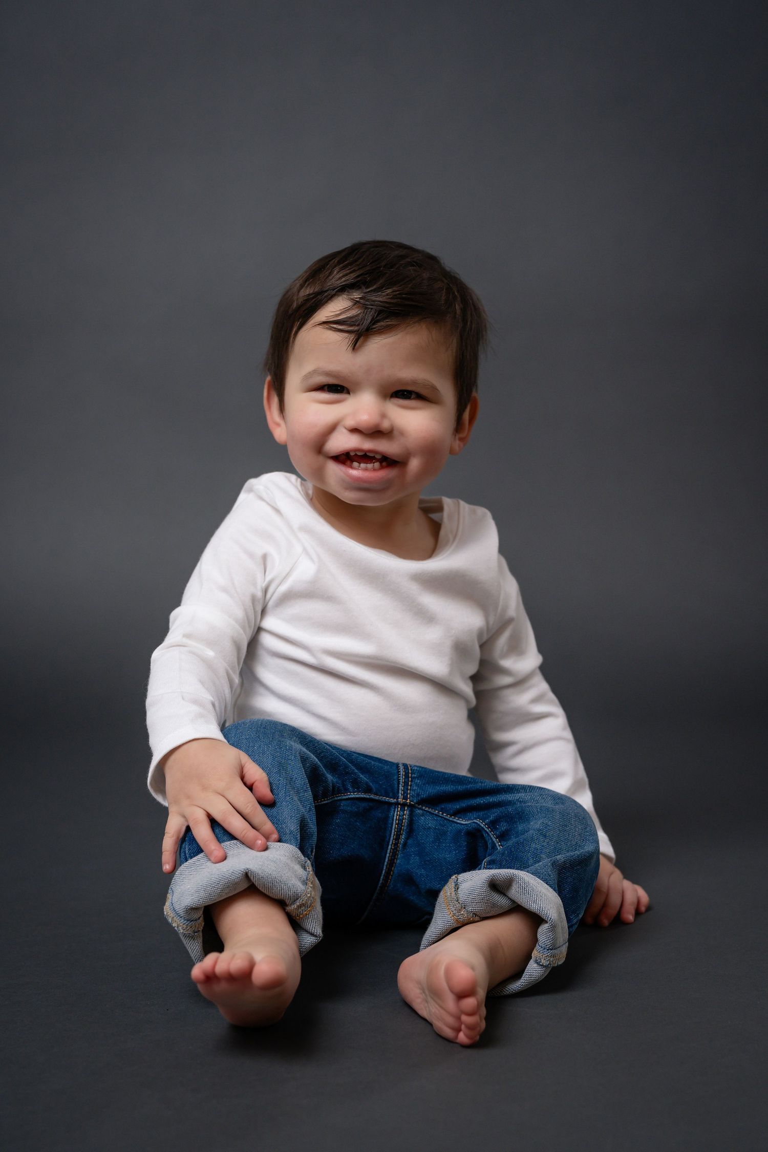 Baby in white top and rolled-up jeans sitting on dark background showing playful expressions.