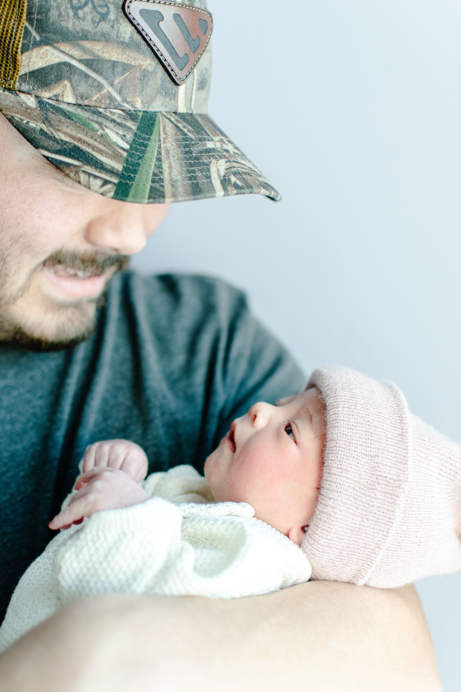 Baby wearing pink hat and white outfit being held by person in camo baseball cap.