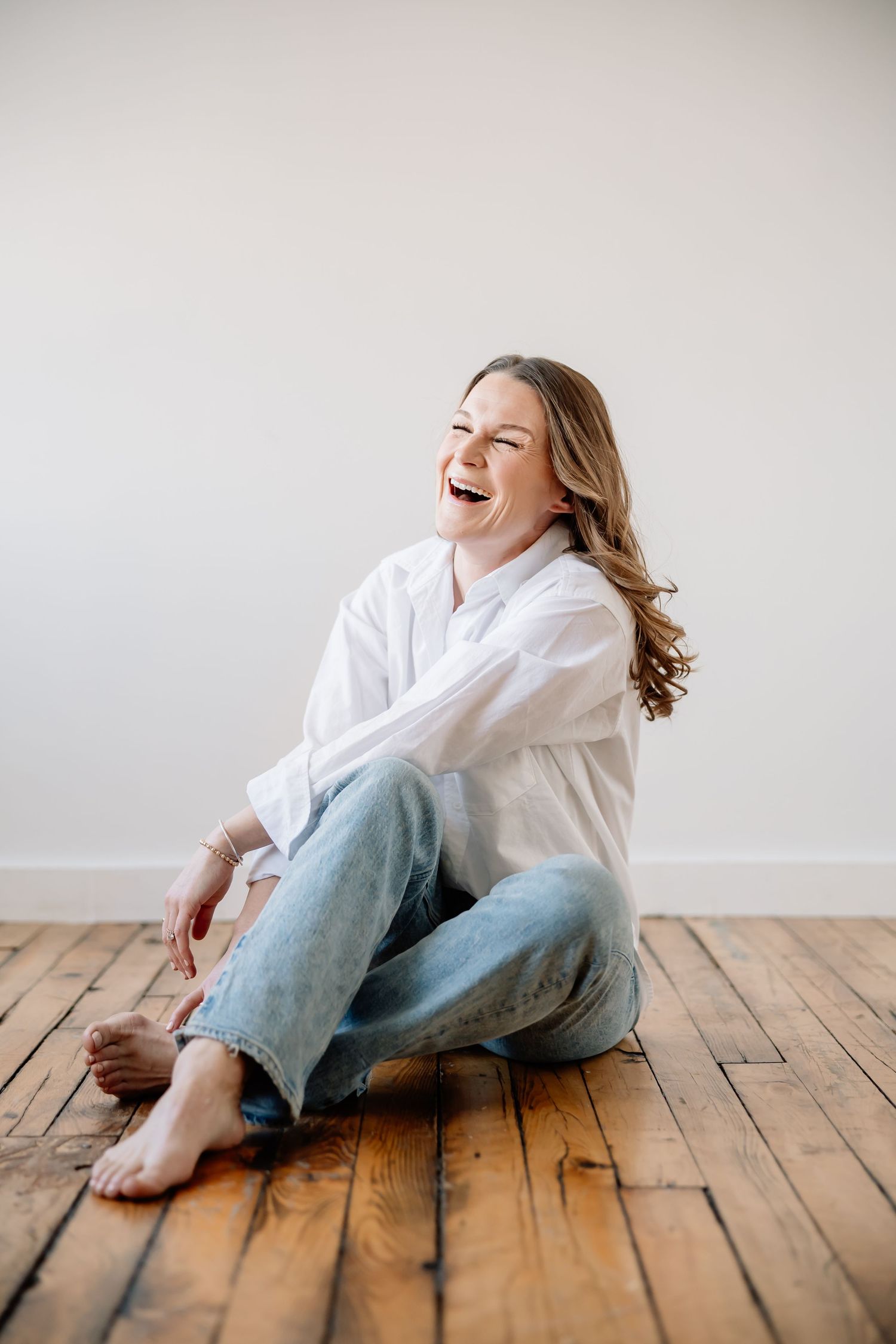 A person in a white shirt and jeans laughs joyfully while sitting casually on a hardwood floor.