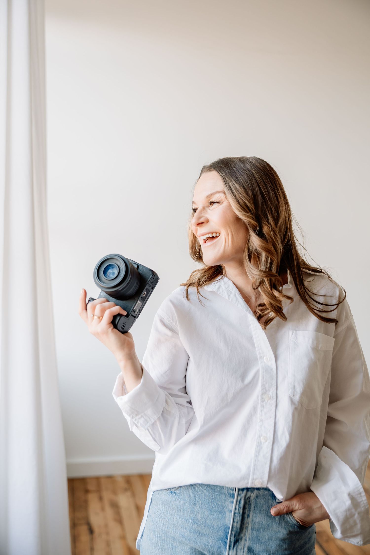 Woman in white shirt and jeans laughing joyfully while holding coffee mug against bright white wall background.