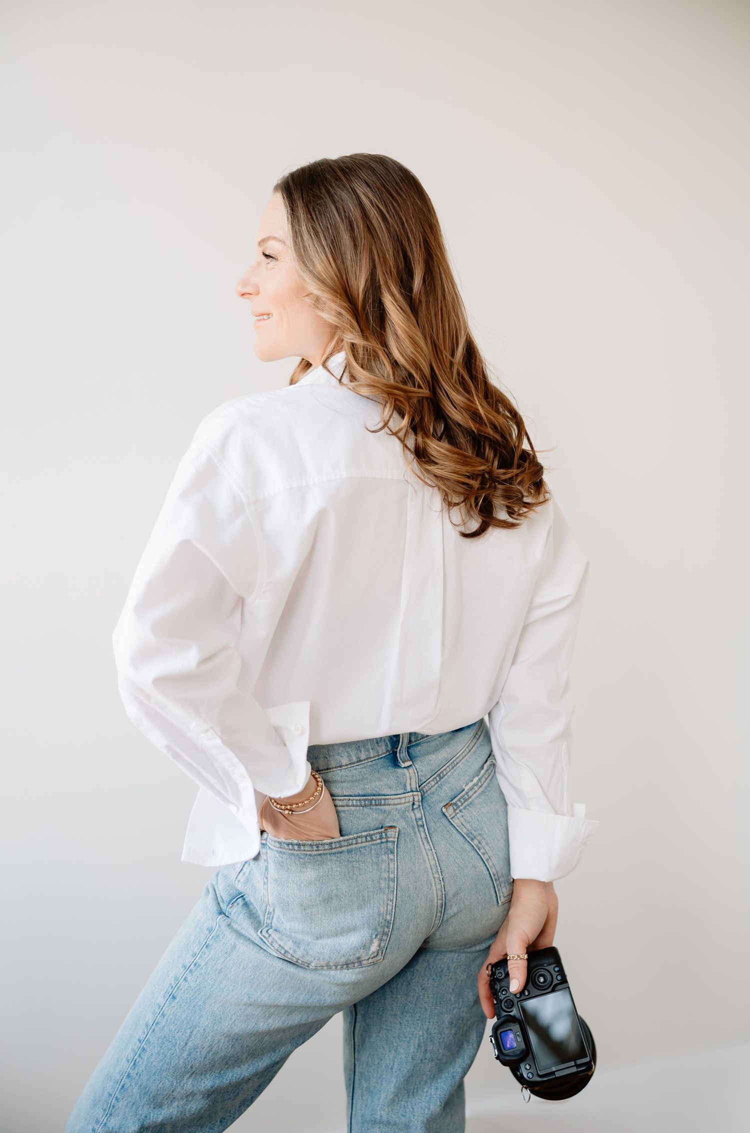 Woman in white blouse and light wash jeans poses against white wall in dynamic fashion editorial style series.