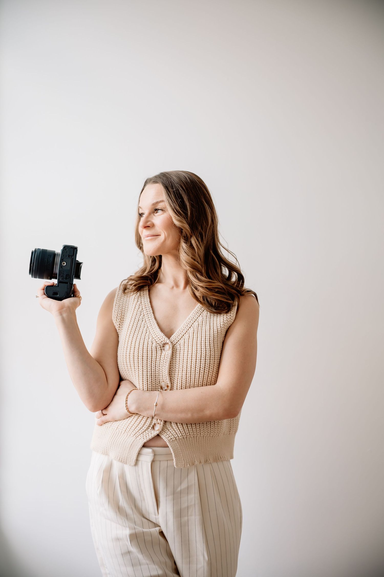Professional photographer in beige dress holds camera while posing against white studio backdrop in three poses.
