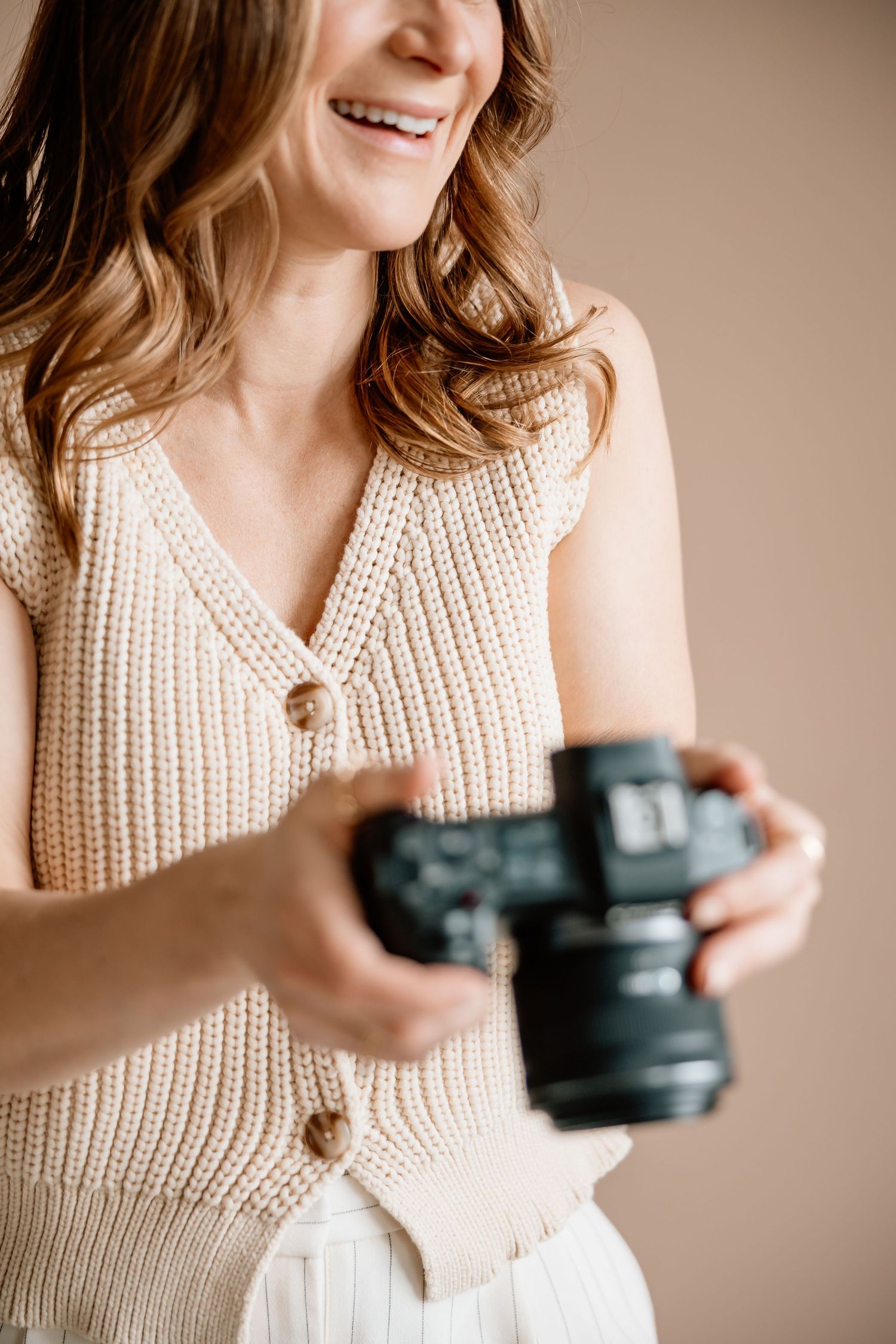 Person wearing cream knit sweater holds black camera while smiling against neutral background.