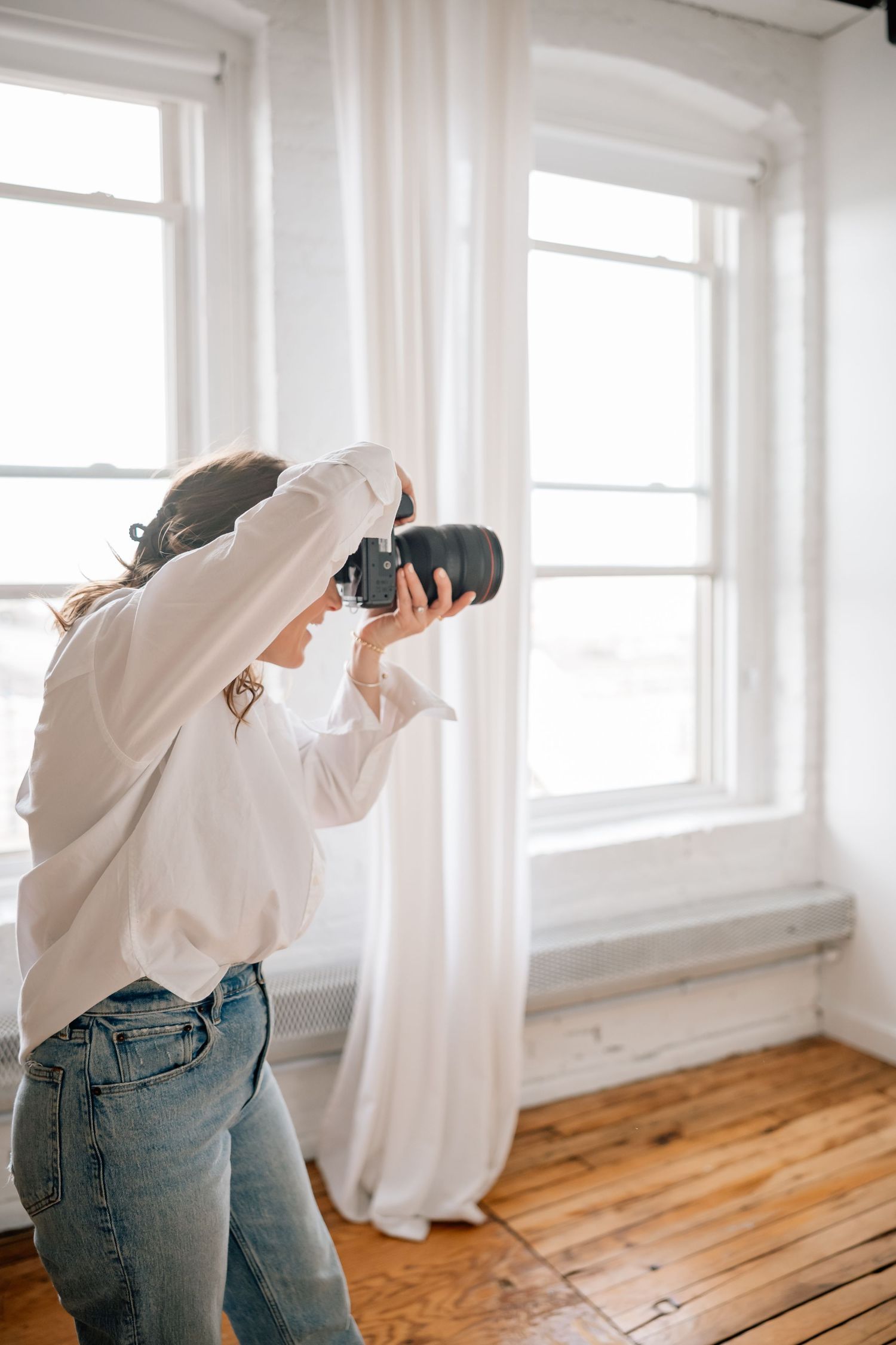 A photographer in jeans and white shirt takes photos in a bright minimalist room with wooden floors and white walls.