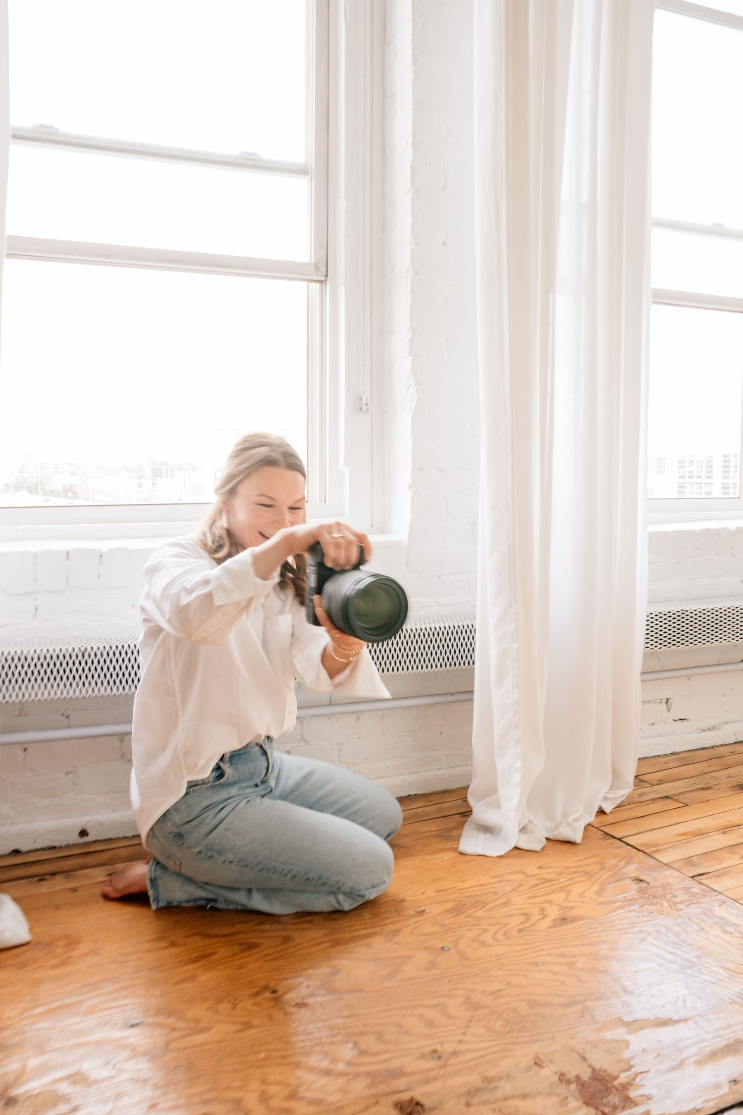 Person in white hoodie and jeans sits on wooden floor near bright windows in minimalist room.