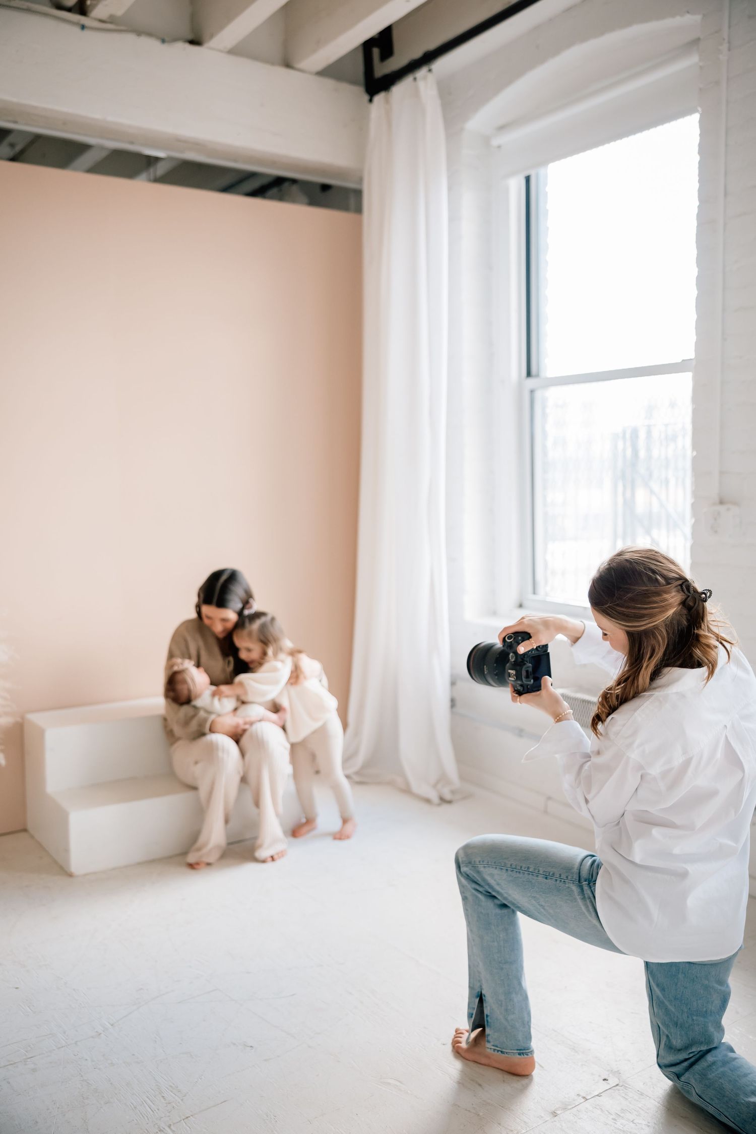Professional photographer in white shirt and jeans taking photos in bright minimalist studio setting.
