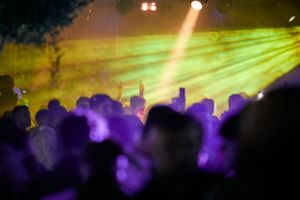 Concert crowd silhouette with bright yellow spotlight beam cutting through purple atmosphere.