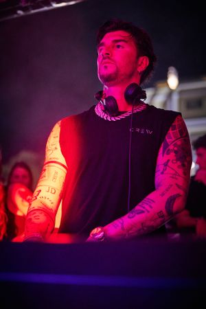 DJ performing at nightclub deck under red and purple lighting.