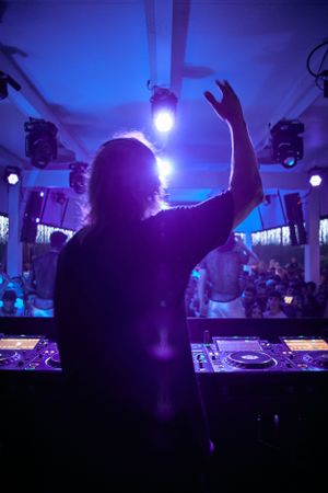 Silhouette of DJ performing against blue-lit club ceiling with hands raised.