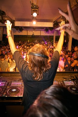 DJ with arms raised performing to packed crowd under vintage lighting fixtures.