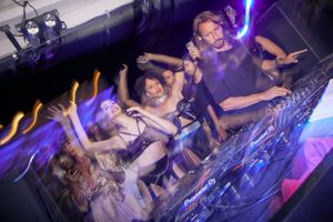 High energy club scene with DJs performing at decks under neon lighting effects.