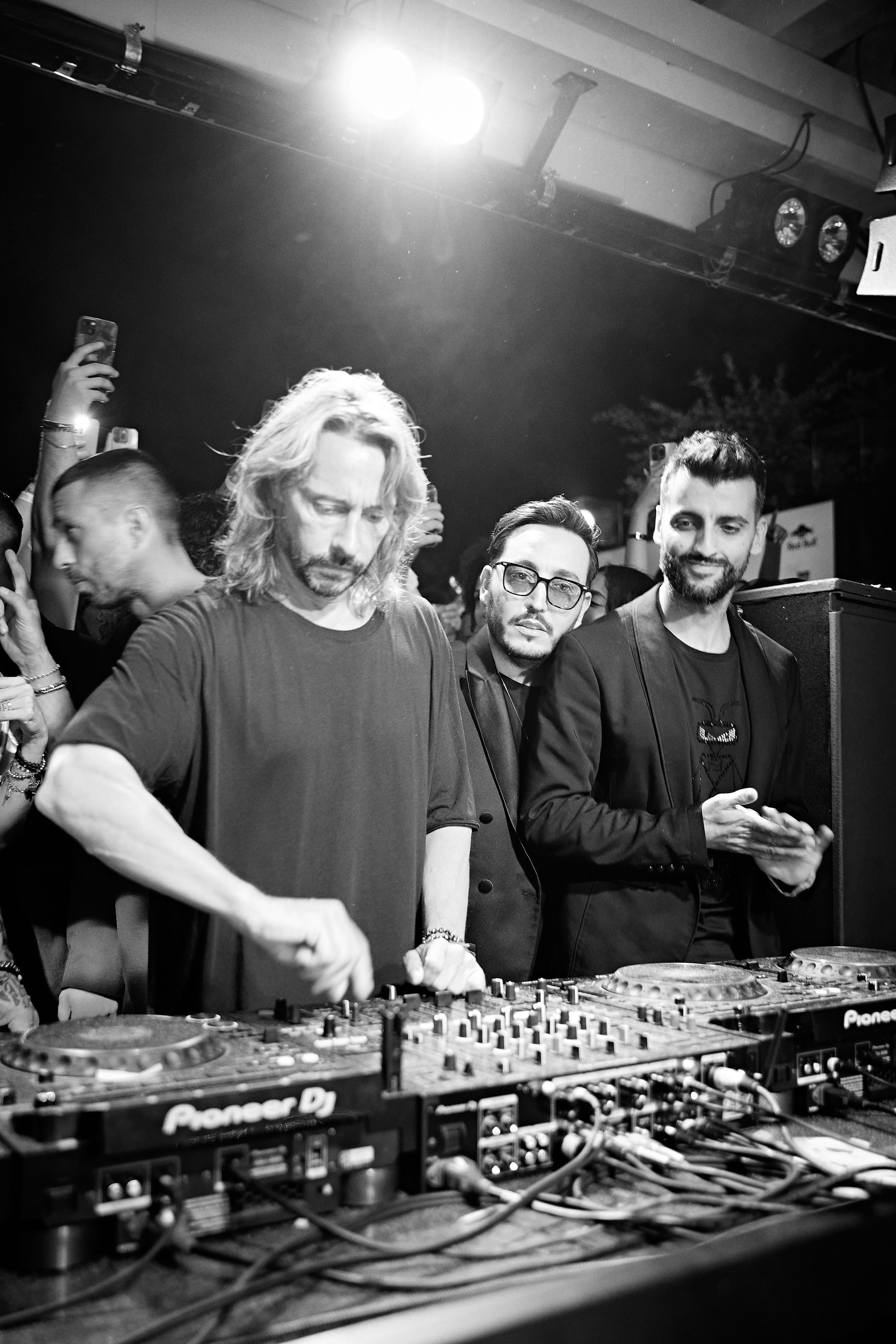Black and white shot of DJ working Pioneer equipment at crowded nightclub.