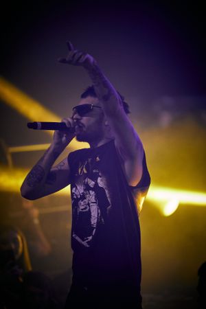 Performer in dark shirt raises arm while singing under yellow stage lights.