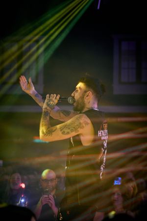 Performer with tattoos singing on stage under colorful lighting during a live concert performance.
