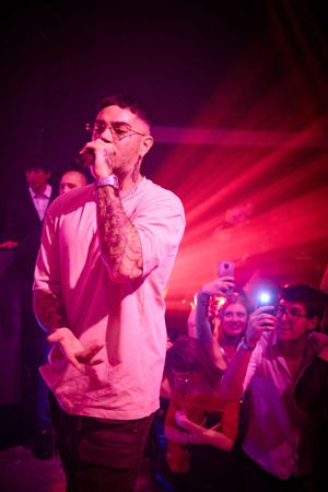 Tattooed performer in white shirt sings on stage with red lighting while audience records on phones.