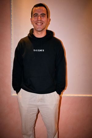 Person in black hoodie and white pants poses against pink wall.