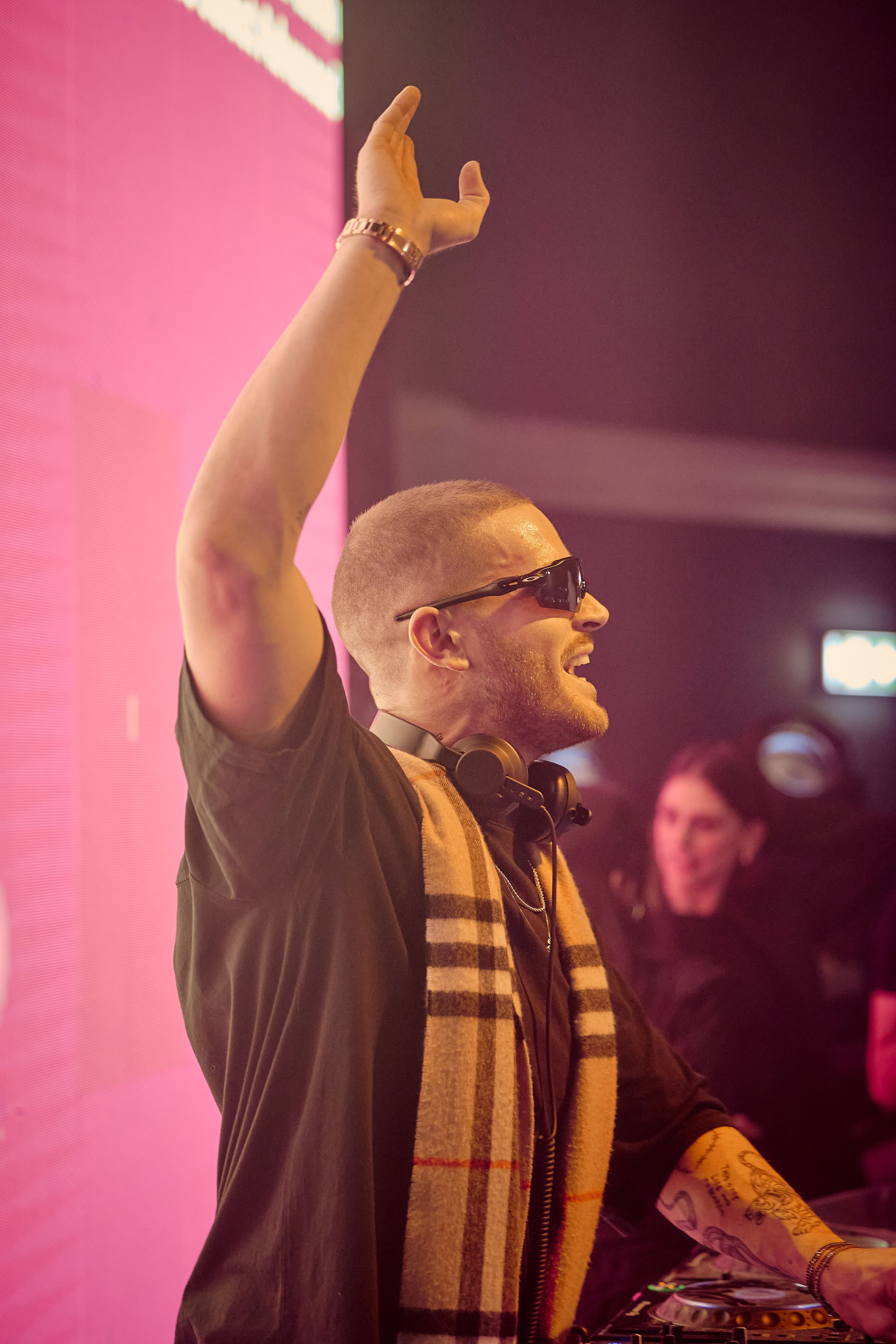 DJ raising arm while performing at decks wearing sunglasses and striped scarf.