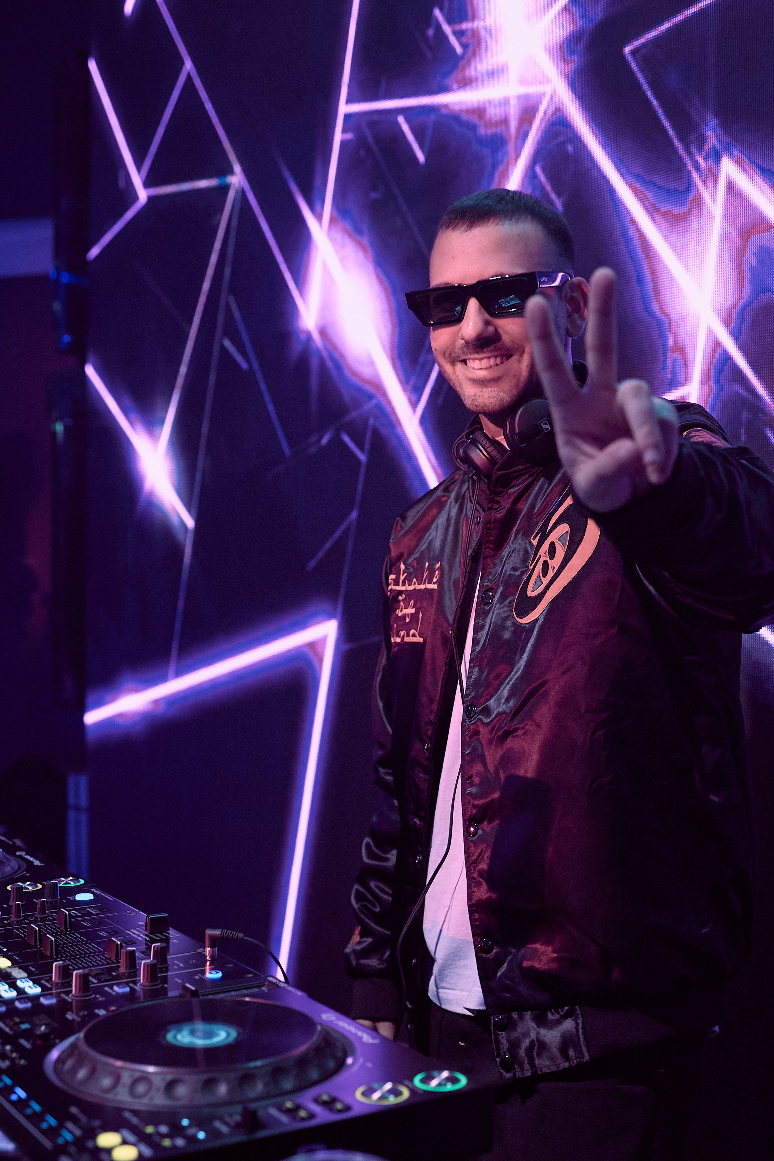DJ performs at nightclub with purple neon geometric lights in background.