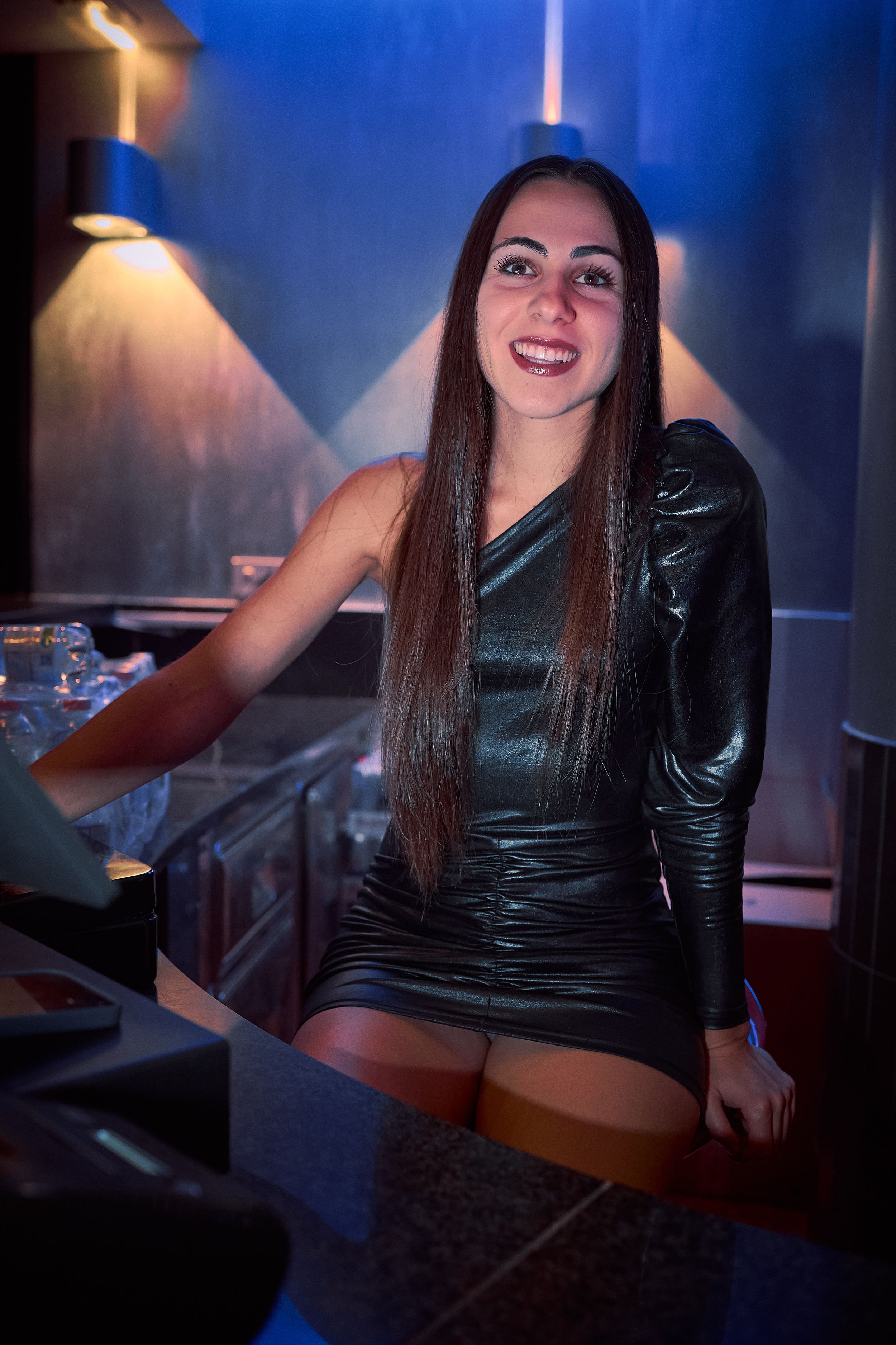 DJ performing at a nightclub's mixing desk wearing a black shiny dress under blue lighting.