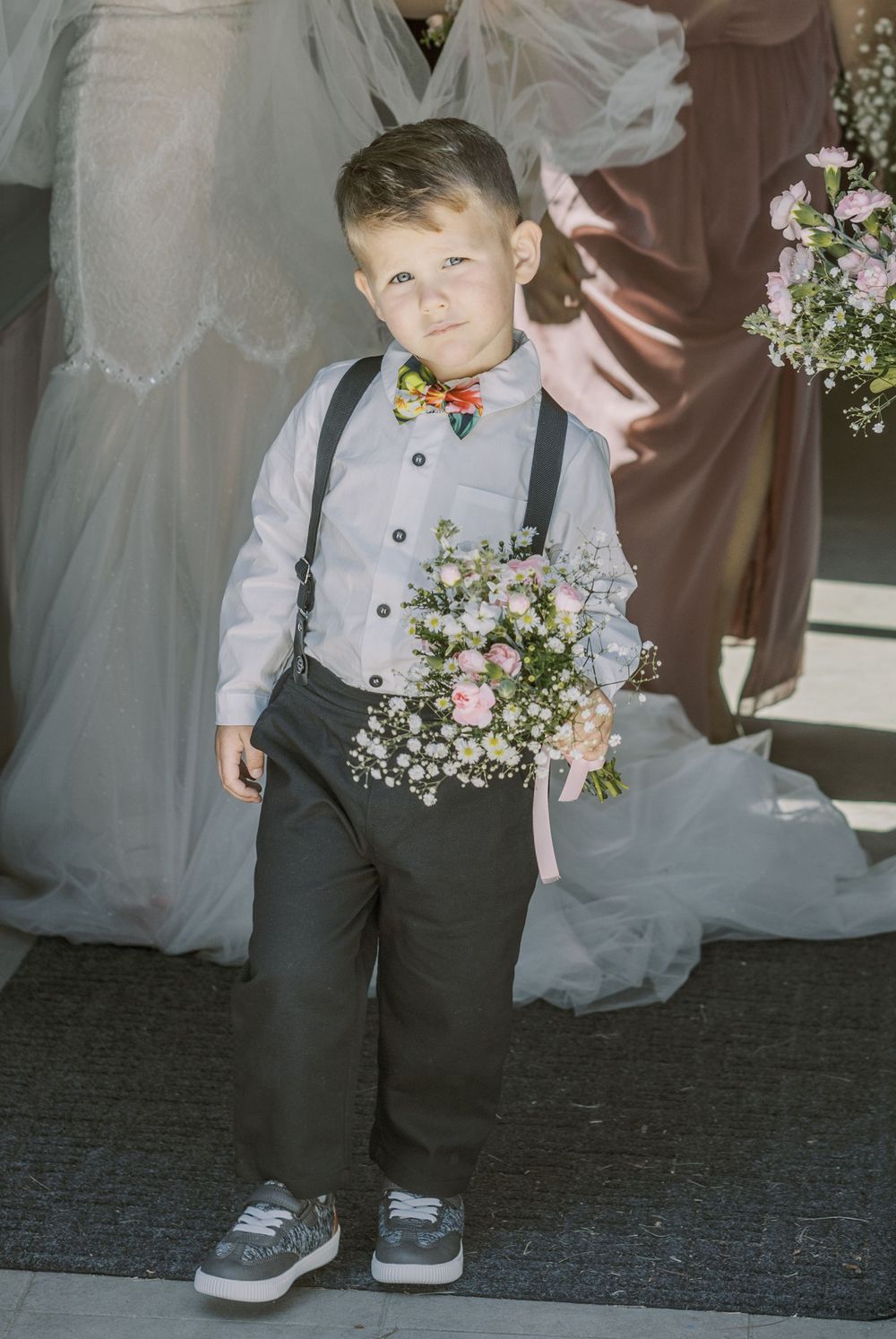 Young ring bearer in formal attire with gray vest walks at wedding ceremony holding delicate flowers.