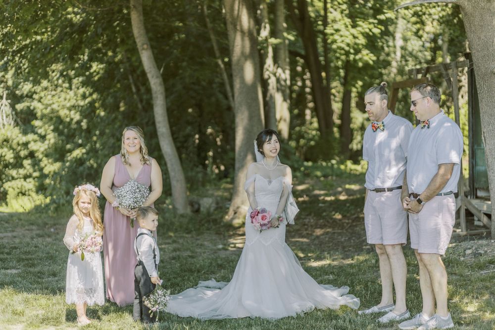 Elegant outdoor wedding ceremony with attendants in light colored dresses against wooded backdrop.