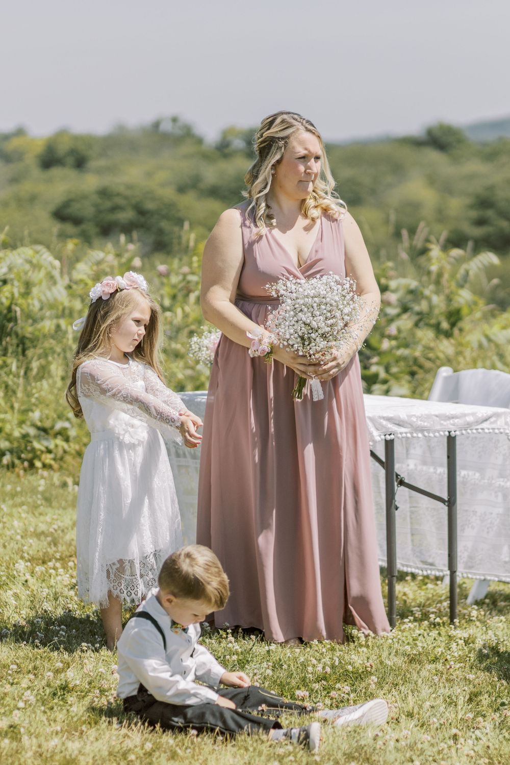 Wedding attendants in blush pink dresses gather around white vintage furniture outdoors.