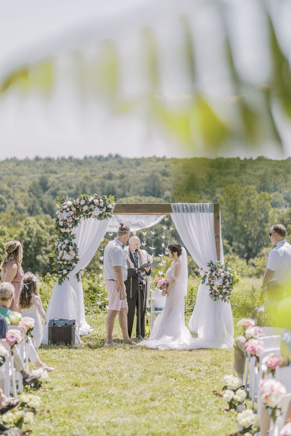 Panoramic view of outdoor wedding setup with white draped canopies and floral decorations on grassy hillside.