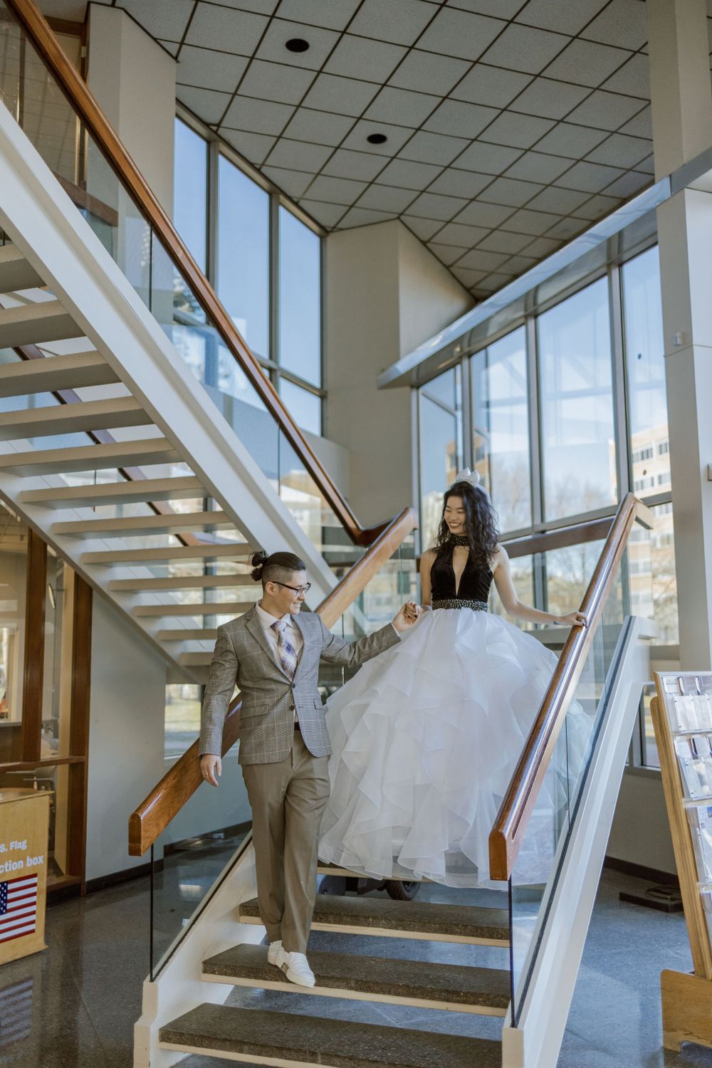 A couple in wedding attire descends a modern staircase with large windows in a bright interior space.