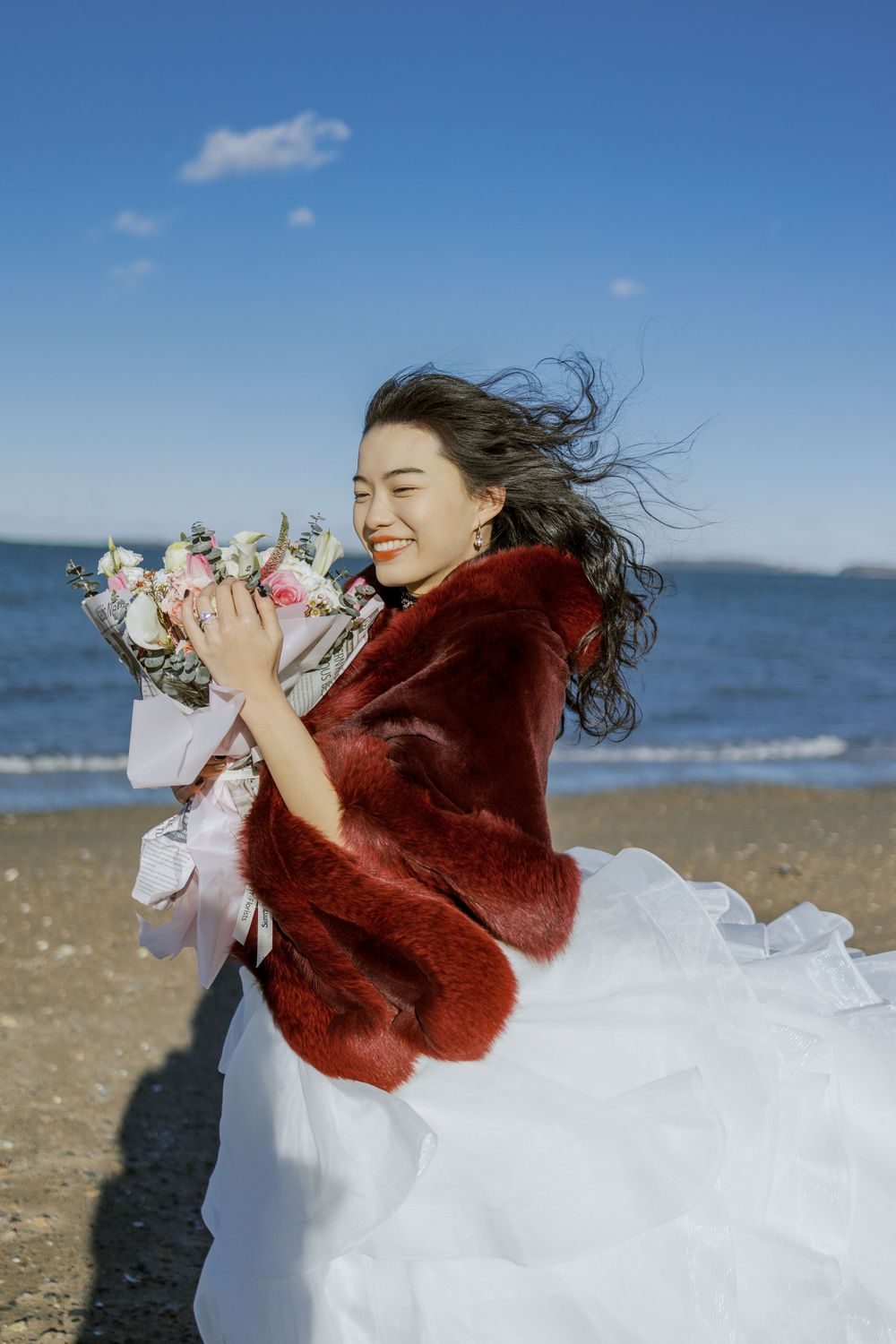 A bride in a white dress and burgundy shawl poses playfully at the beach on a sunny day.