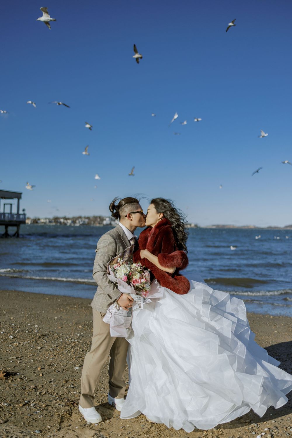 A couple embraces on a beach with seagulls flying overhead against a bright blue sky.