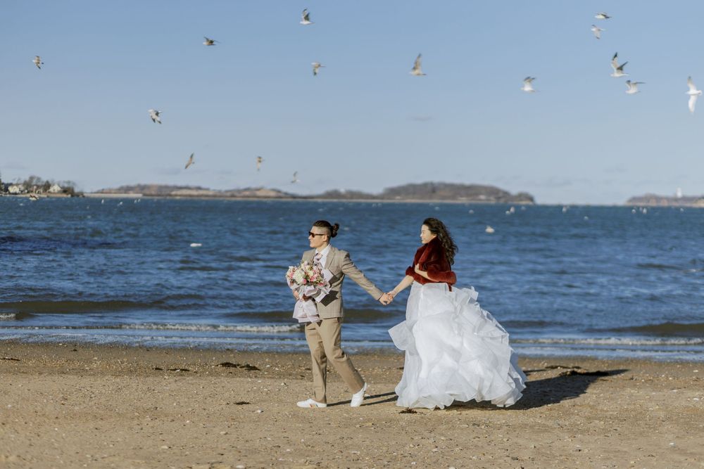 A couple shares a playful moment on a beach with seagulls flying overhead.