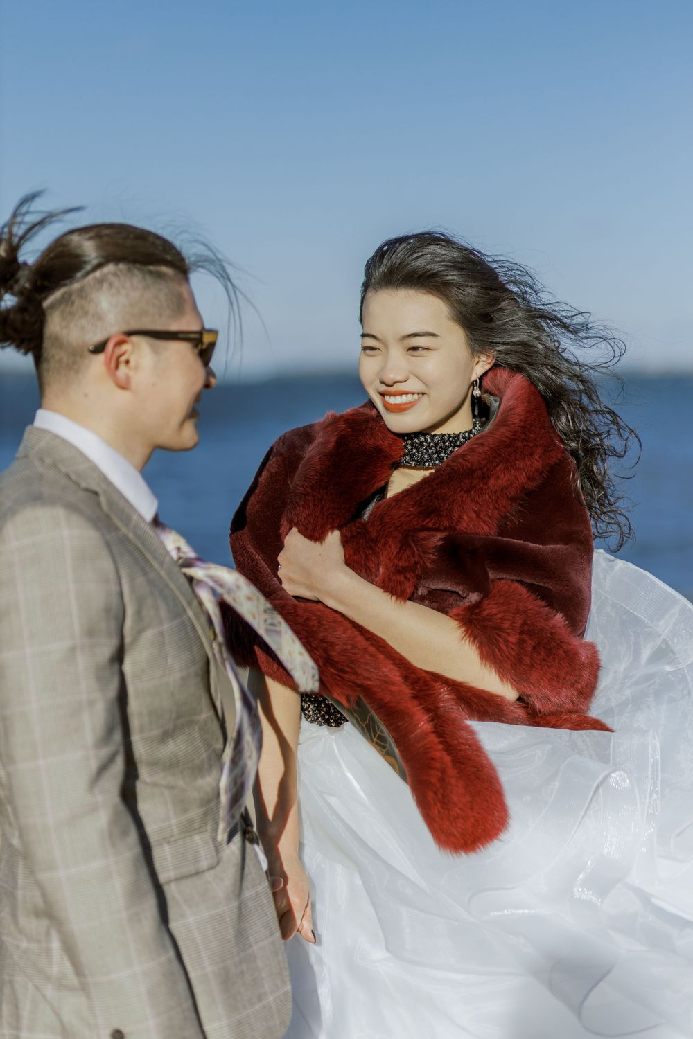 A wedding couple shares a joyful moment by the ocean, with the bride wearing a red fur shawl over her white dress.