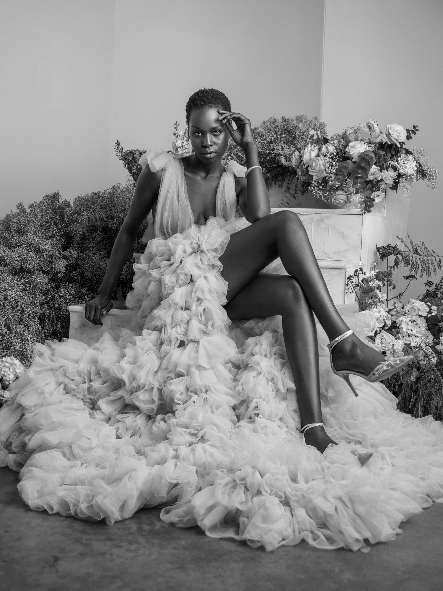Black and white photo of a woman in a ruffled tulle gown seated among flowers.