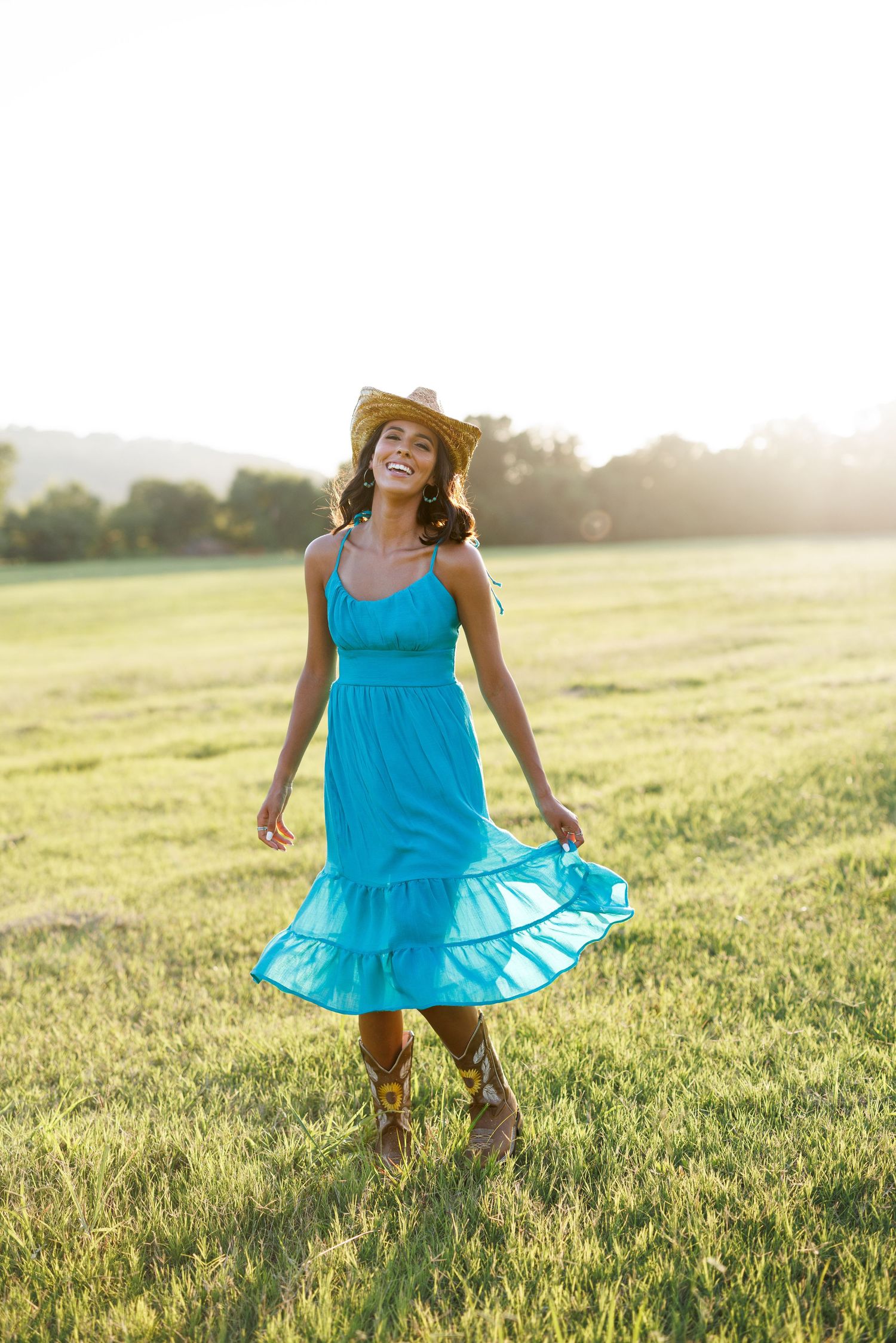 Woman in bright blue dress and cowboy boots twirls playfully in a sunlit field.