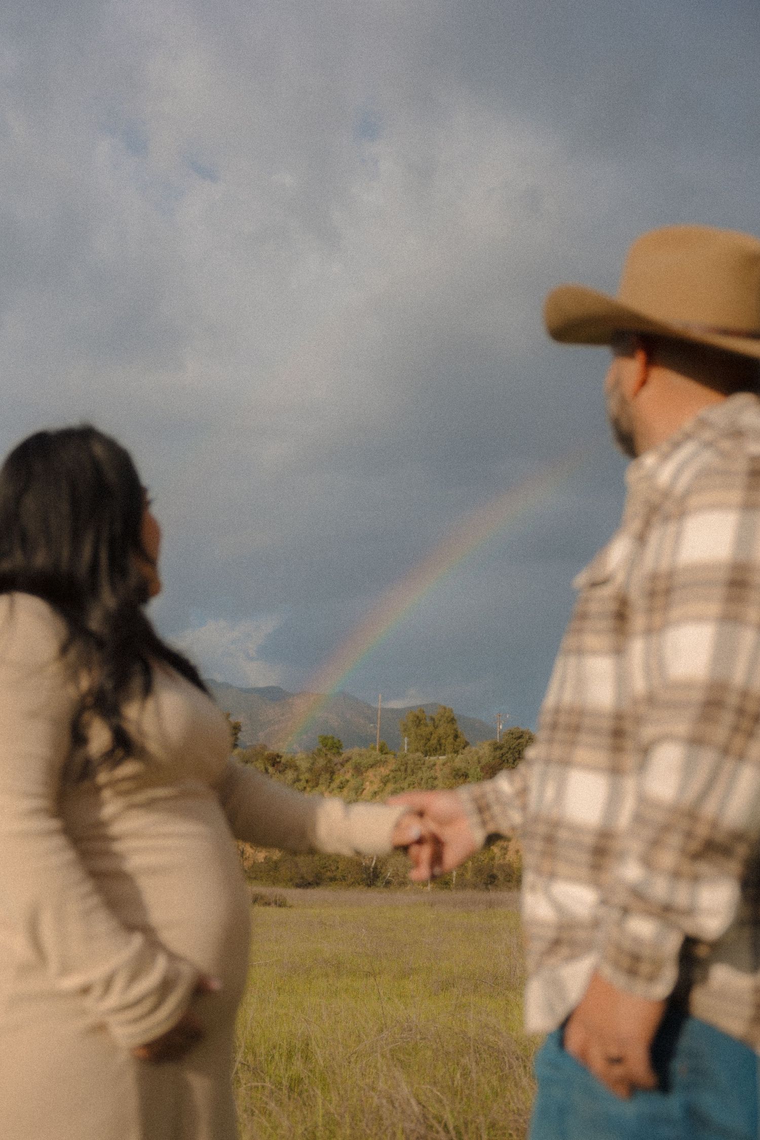 A couple holds hands while gazing at a rainbow arching across a cloudy rural landscape.