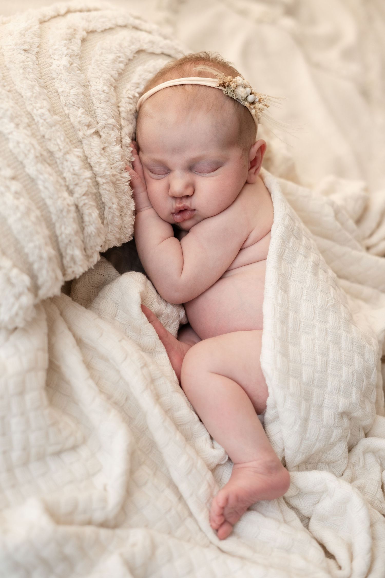 Sleeping newborn baby girl wrapped in cream blanket wearing floral headband.
