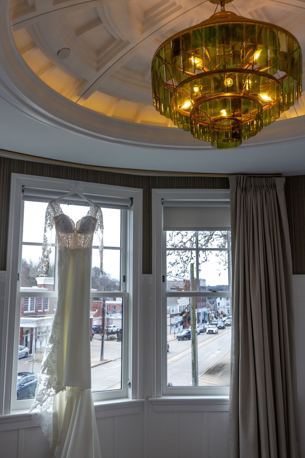 Lace wedding dress hanging in a bay window beneath a green glass chandelier with town view outside.