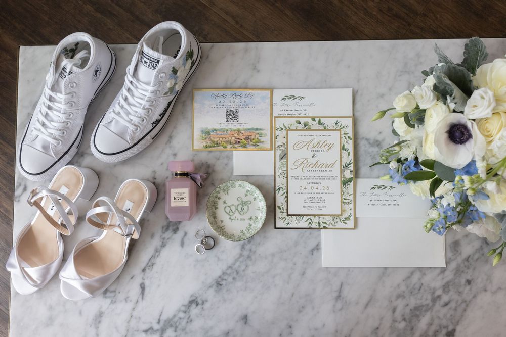 Bridal flat lay featuring white Converse, heels, floral invitations, perfume, and blue and white bouquet.