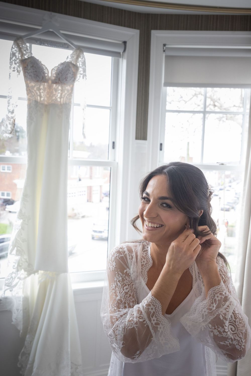 Smiling bride in white lace robe putting on earrings with wedding dress hanging in window behind her.