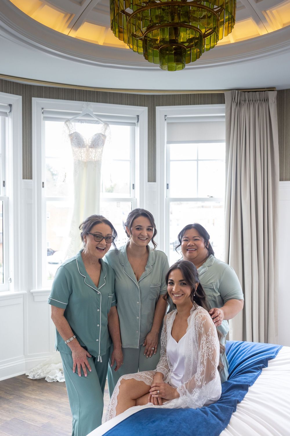 Bride sitting on bed with three bridesmaids in sage green pajamas posing in elegant hotel suite.