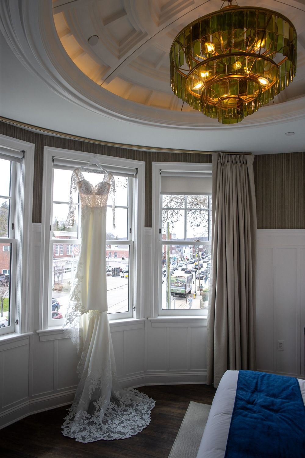 Elegant wedding dress hanging in a bay window room with a green chandelier and city street view below.