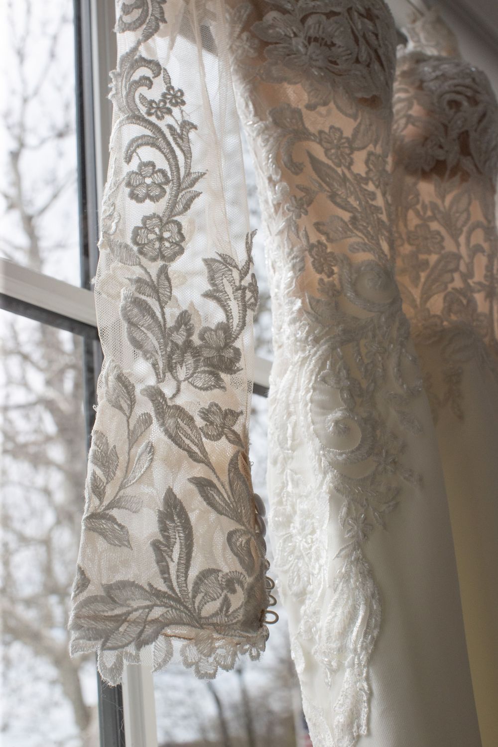 Close-up of intricate floral lace detail on a bridal gown hanging by a frosted window.