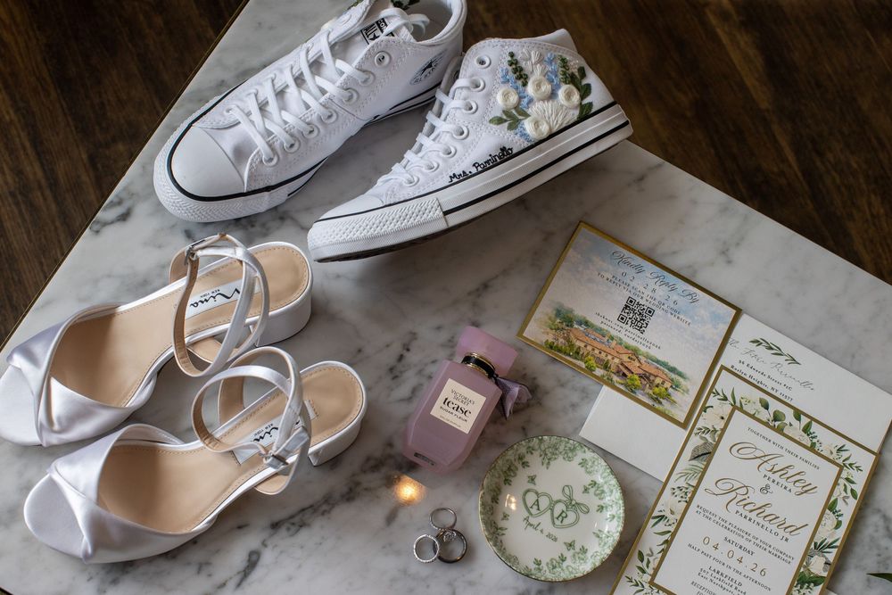 Wedding flat lay with custom floral Converse, silver heels, invitations, rings, and pink perfume.