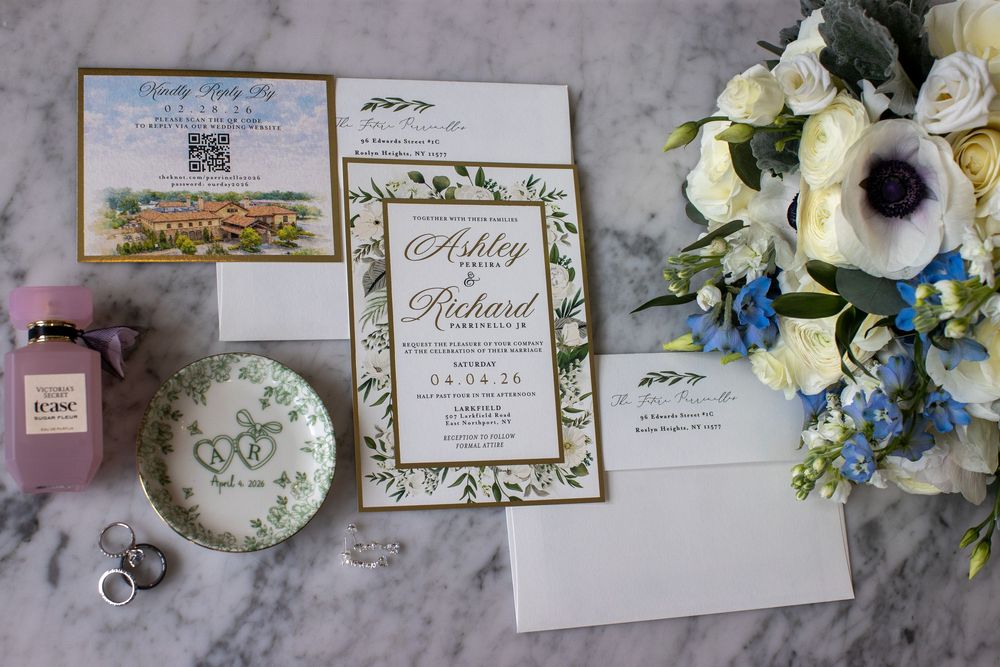 Wedding stationery flat lay with floral invitations, bouquet, rings, and pink perfume bottle.