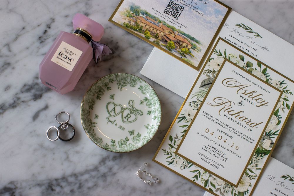 Close-up of elegant gold-bordered wedding invitations with monogram dish and pink perfume bottle.