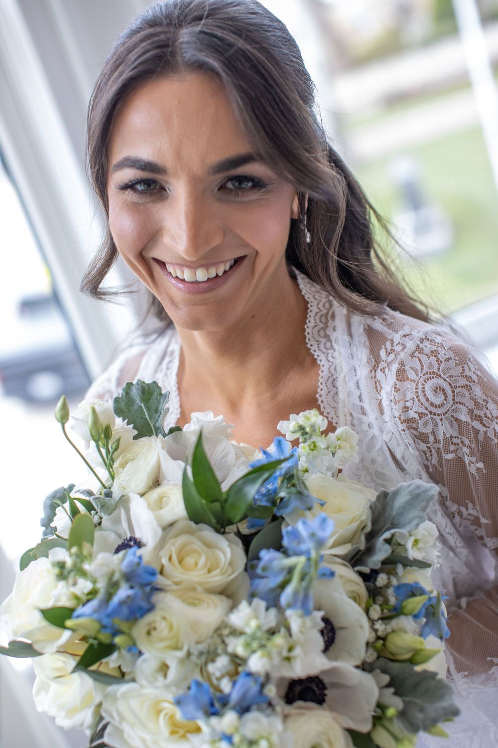 Smiling bride in lace robe holding white and blue floral bouquet with anemones and roses.
