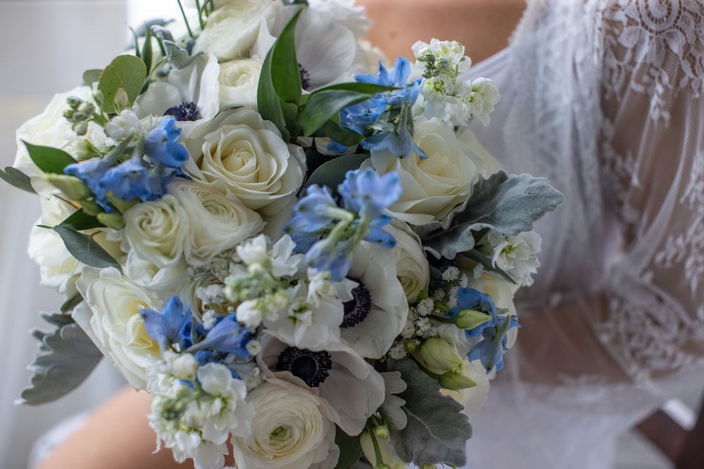 Bridal bouquet featuring white roses, anemones, blue delphinium, and dusty miller greenery.