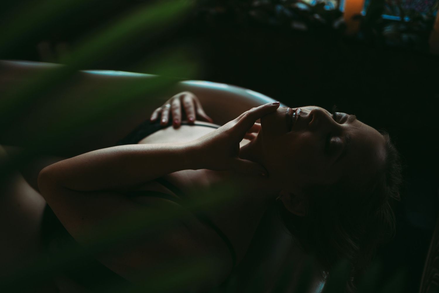 Woman in black lingerie, lying in a vintage bathtub, with soft lighting and an intimate, sultry expression, captured through a plant