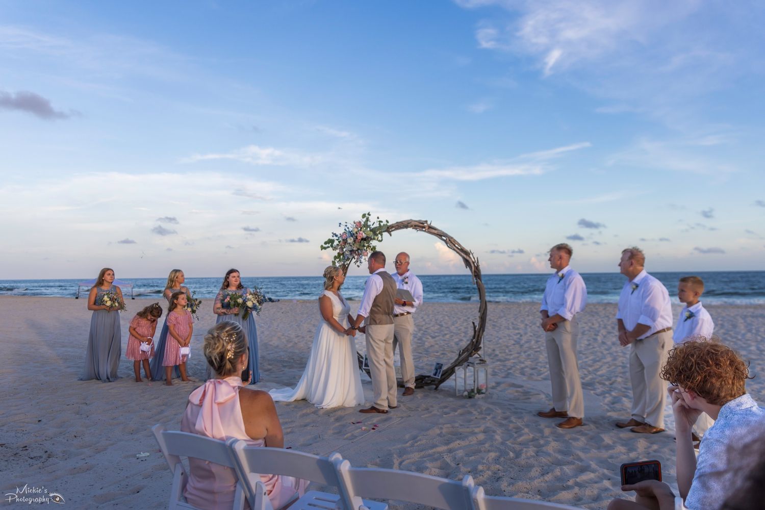 Beach wedding ceremony with circular floral arch and guests gathered on sand.